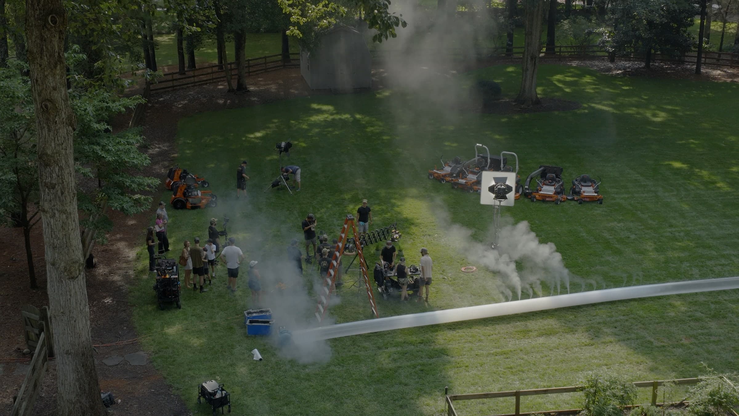A drone plate looking down on the unit working a lawn: crew gathered around the camera and lighting rigs, smoke drifting across the grass and mowers staged at the treeline