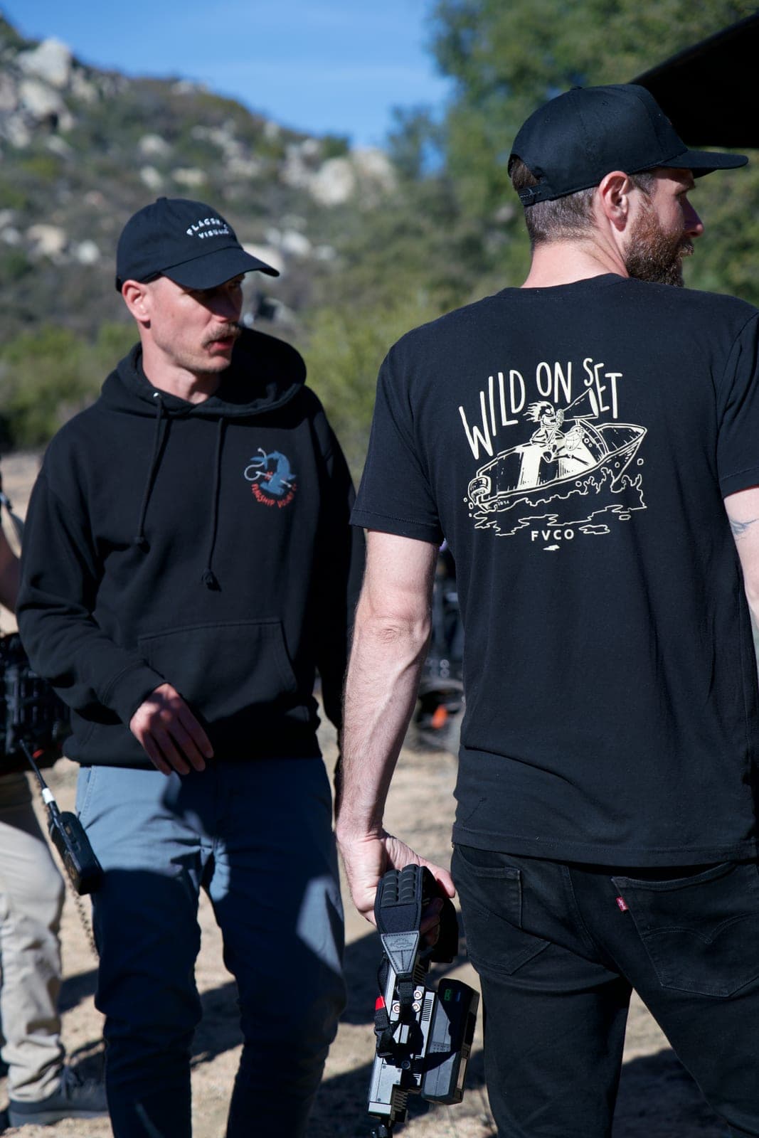 Two of the crew walking back between takes, one carrying a camera body, hills behind them