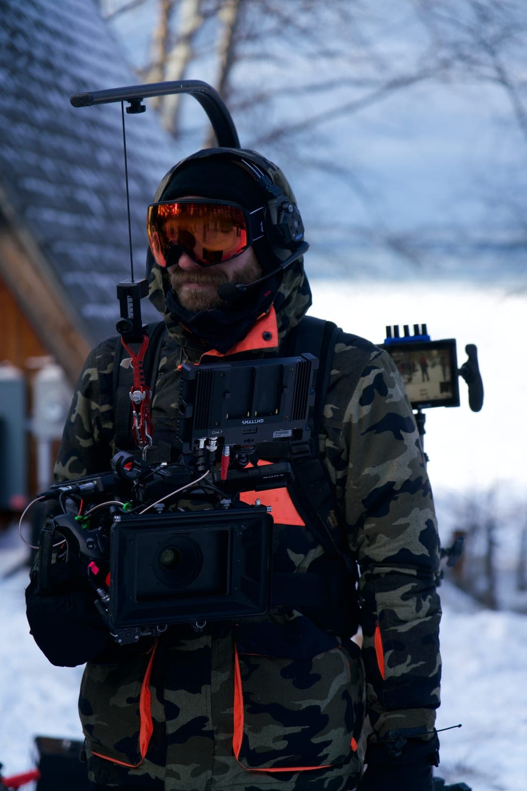 A camera operator in insulated camouflage and goggles holding a rigged cinema camera in deep snow