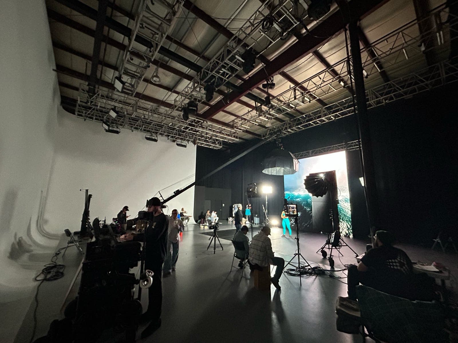 A sound stage seen from the cyc wall: lighting grid overhead, a lit set at the far end and the crew working in the dark floor between